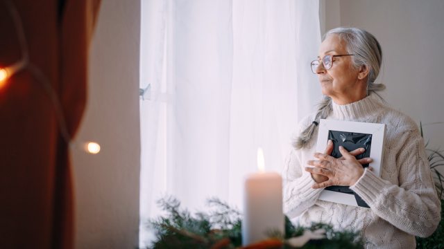 A woman holds a photo of a loved one who has passed close to her chest. She sits near a window and a lighted candle.